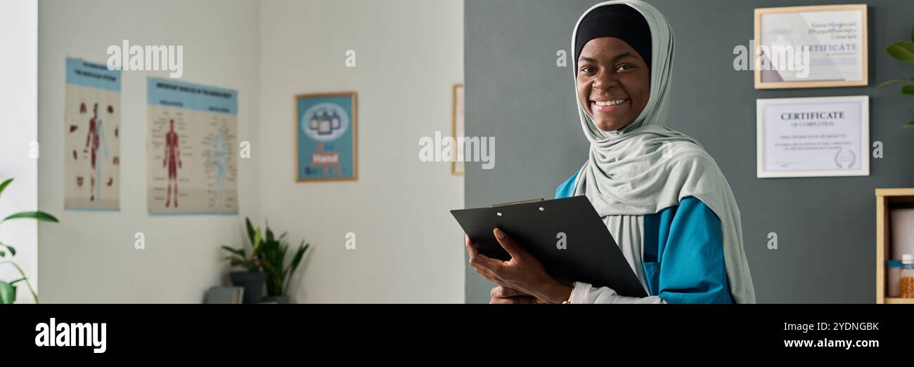 Giovane donna musulmana afroamericana in piedi in ambiente sanitario sorridente calorosamente mentre tiene gli appunti e indossa un abbigliamento professionale con certificati sul muro in background Foto Stock