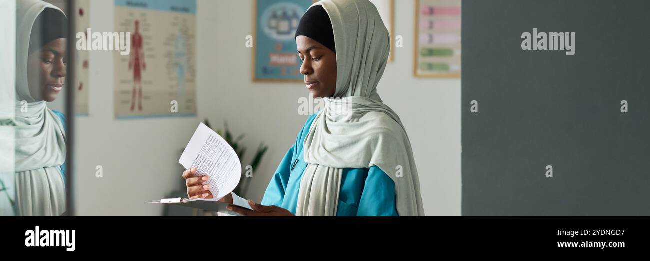 Vista laterale di una donna musulmana africana che indossa un hijab, legge documenti in uno studio medico, con una cartella anatomica visibile sullo sfondo Foto Stock