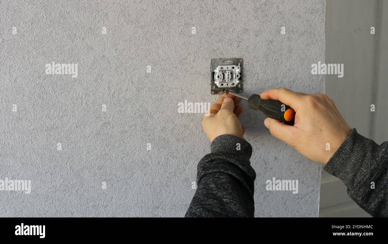 Un lavoratore fissa una vite a una presa o a un interruttore luce con un cacciavite su uno sfondo grigio Foto Stock
