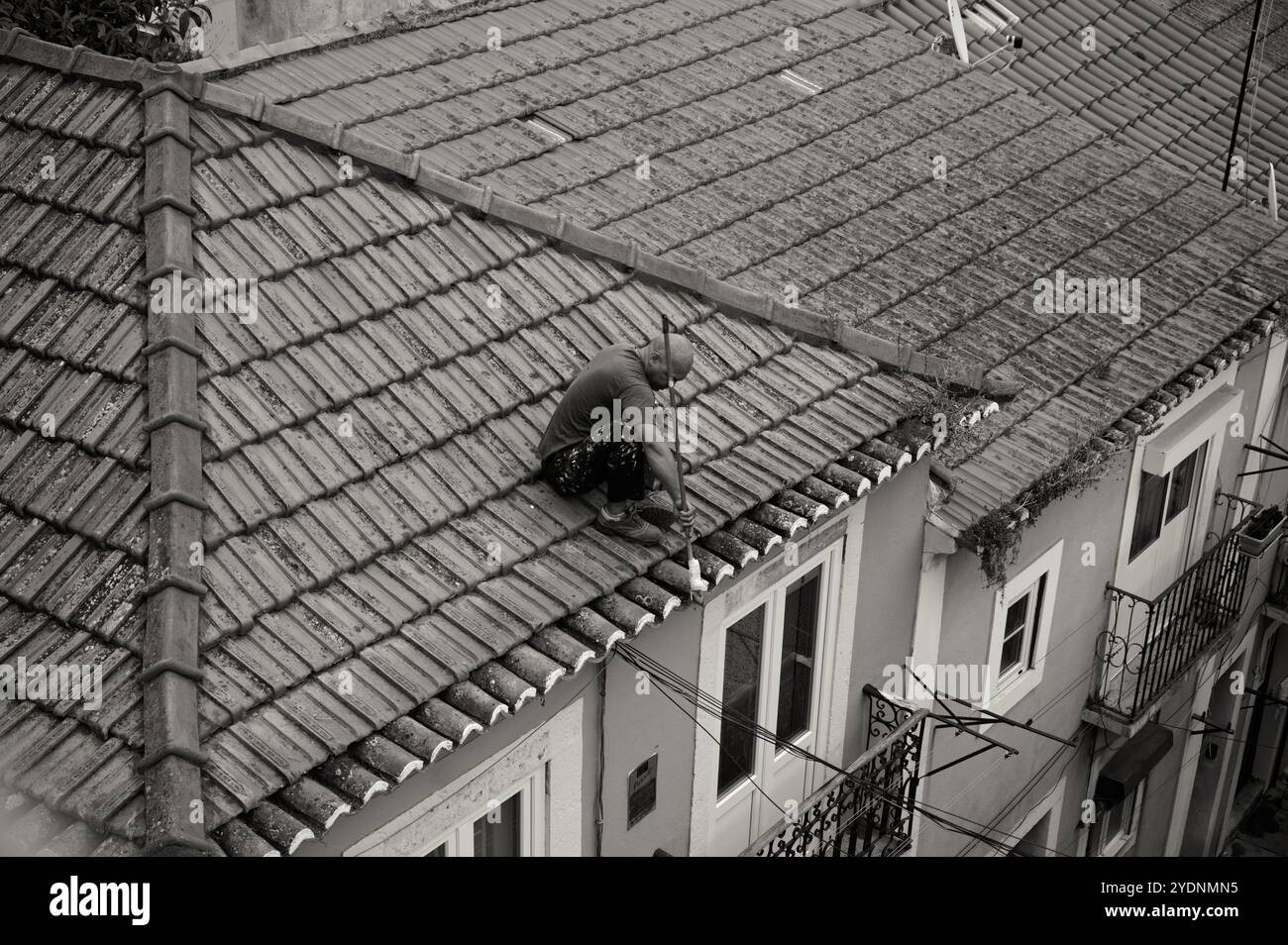 Uomo che pulisce un tetto in piastrelle ad Alfama, si concentra sulla manutenzione in alto sulla strada in b&W Foto Stock