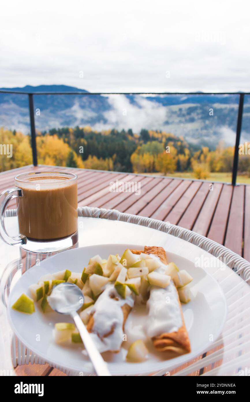 Pancake con frutta tritata e tazza di caffè sullo sfondo mozzafiato del paesaggio autunnale. Colazione in montagna. Colazione su una terrazza in legno in montagna Foto Stock