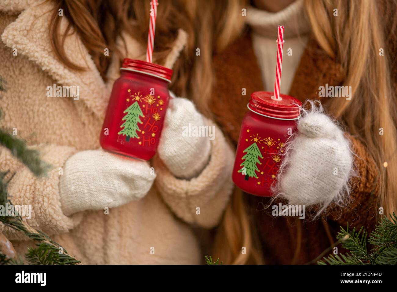 Due amici che indossano comodi maglioni e guanti tengono gioiosamente in mano tazze rosse adornate con disegni di alberi di Natale durante una celebrazione invernale. Foto Stock