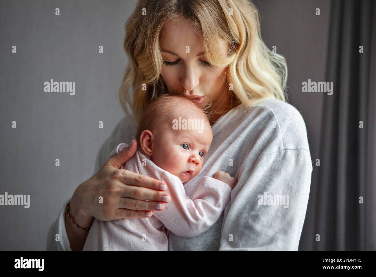 Bella giovane madre bionda che bacia il bambino, vestita con abiti leggeri al coperto. Mamma e neonato su sfondo grigio, tenero legame amorevole tra mamma Foto Stock