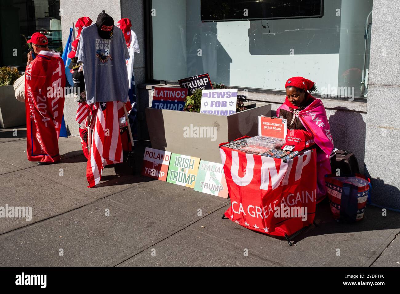 New York, New York, Stati Uniti. 27 ottobre 2024. Folle di sostenitori di Donald Trump, molti con berretti e camicie marchiati MAGA o Trump, si riuniscono fuori dal Madison Square Garden prima di un raduno serale. Un venditore nero offre cartelli che leggono ebrei, irlandesi e molte altre nazionalità per Trump siede al sole sulla 8th Avenue. Crediti: Ed Lefkowicz/Alamy Live News Foto Stock