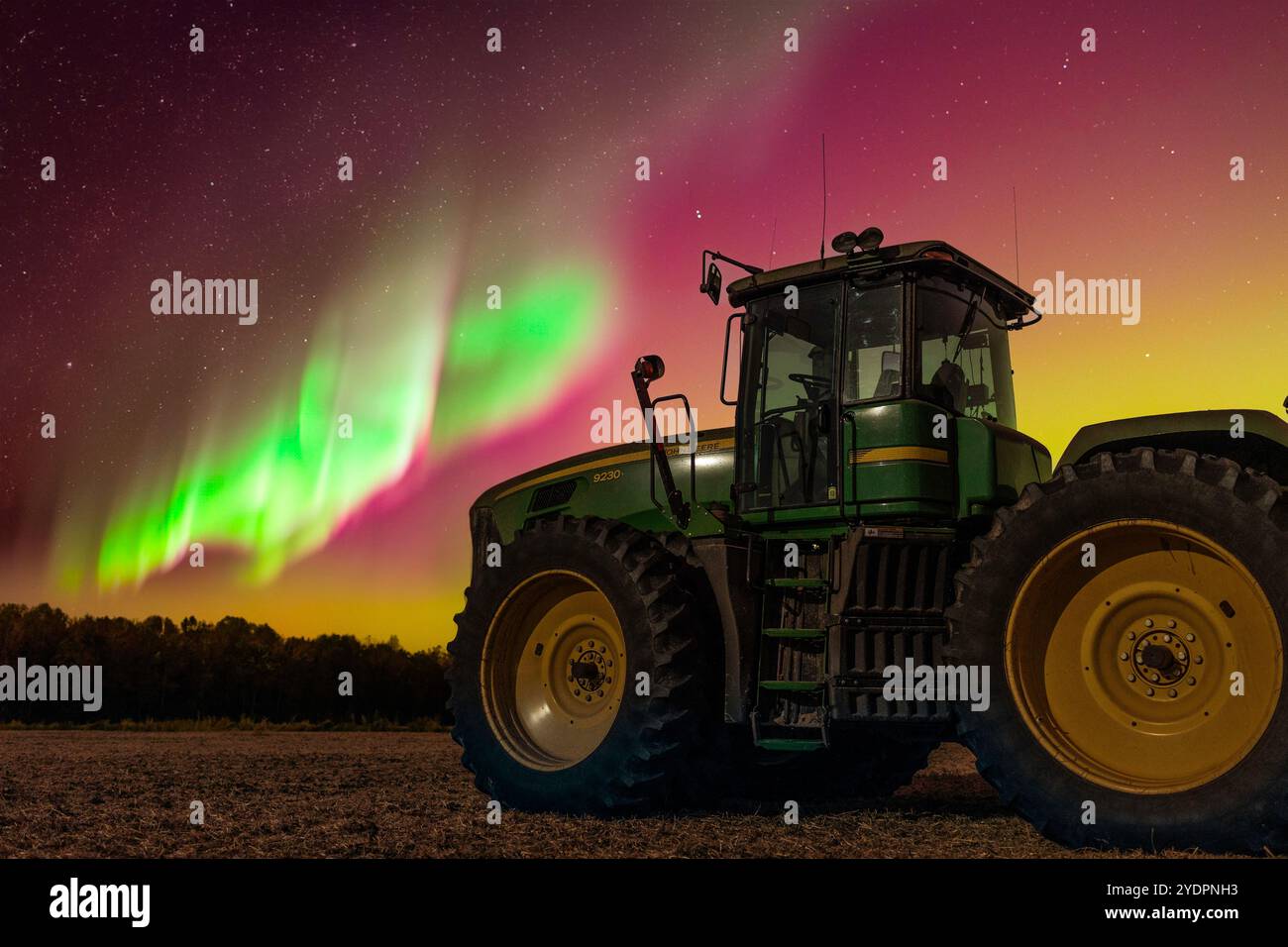 Un trattore articolato si trova in un campo sotto i brillanti nastri verdi e magenta dell'aurora. Il campo di fango raccolto è sullo sfondo. Foto Stock