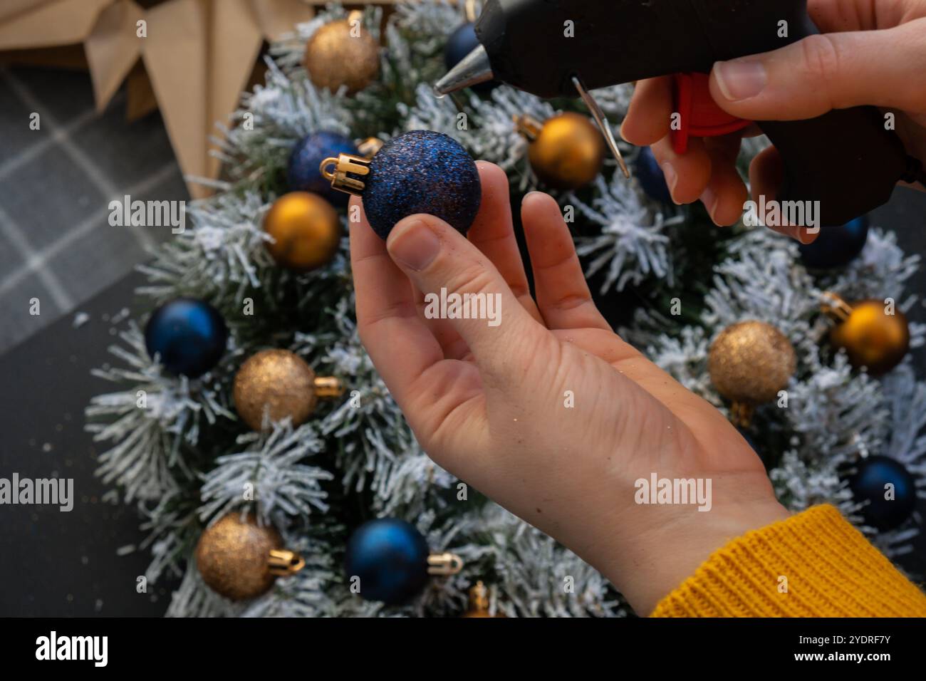 Artigiano con colla calda istruzioni passo dopo passo per realizzare una corona di Natale fatta a mano con decorazioni natalizie nei colori blu e giallo. Mani femminili che creano decorazioni artigianali fatte in casa. Festa di Capodanno. Vacanze invernali fase 2 Foto Stock