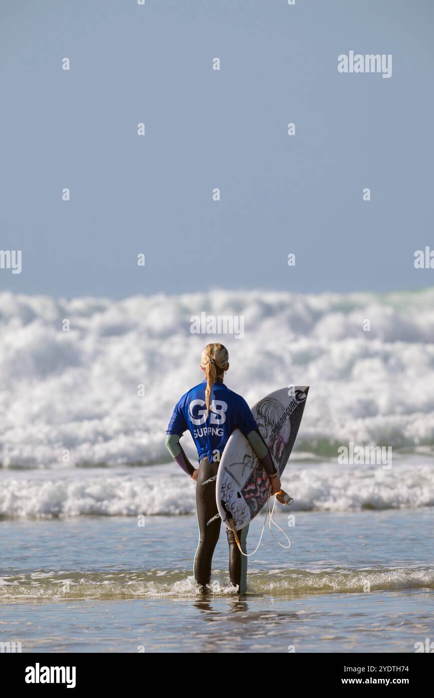 Una surfista con una maglietta GB Surfing sta in acqua, tenendo la tavola, contemplando le onde prima di uscire per fare surf. Foto Stock