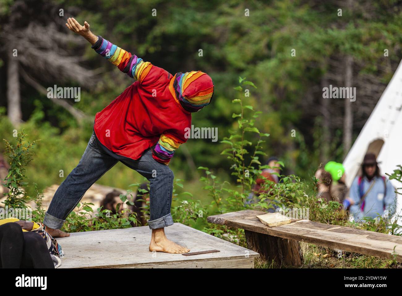 Un istruttore di yoga indossando abiti colorati è visto che spazia da dietro, in piedi nudi in un campeggio durante un raduno di persone spirituali Foto Stock