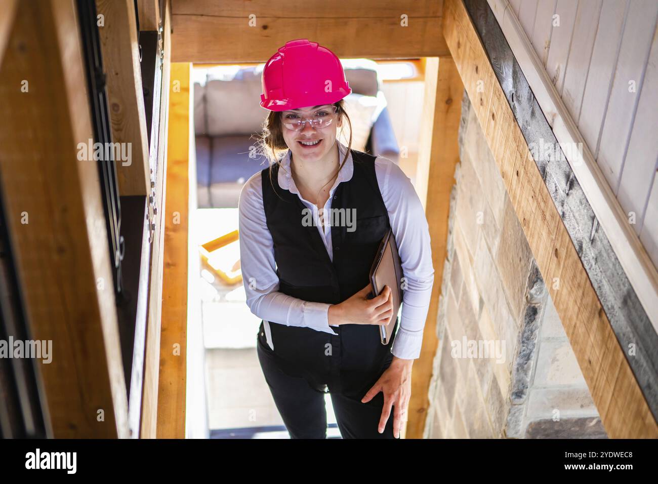 Giovane architetto che sorride sulle scale. Ingegnere civile donna in piedi in cantiere indossando elmetto di sicurezza rosa e tenendo in mano un appunti Foto Stock