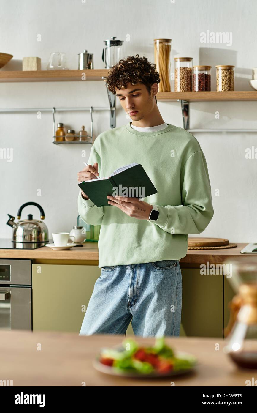 In una cucina contemporanea, un bel giovane scrive diligentemente in un quaderno, circondato da strumenti culinari. Foto Stock