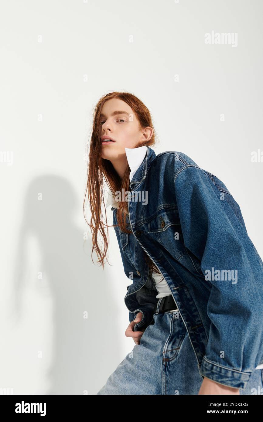 Un ragazzo adolescente sfoggia i suoi lunghi capelli rossi e il suo abito in denim alla moda in un luminoso ambiente da studio. Foto Stock