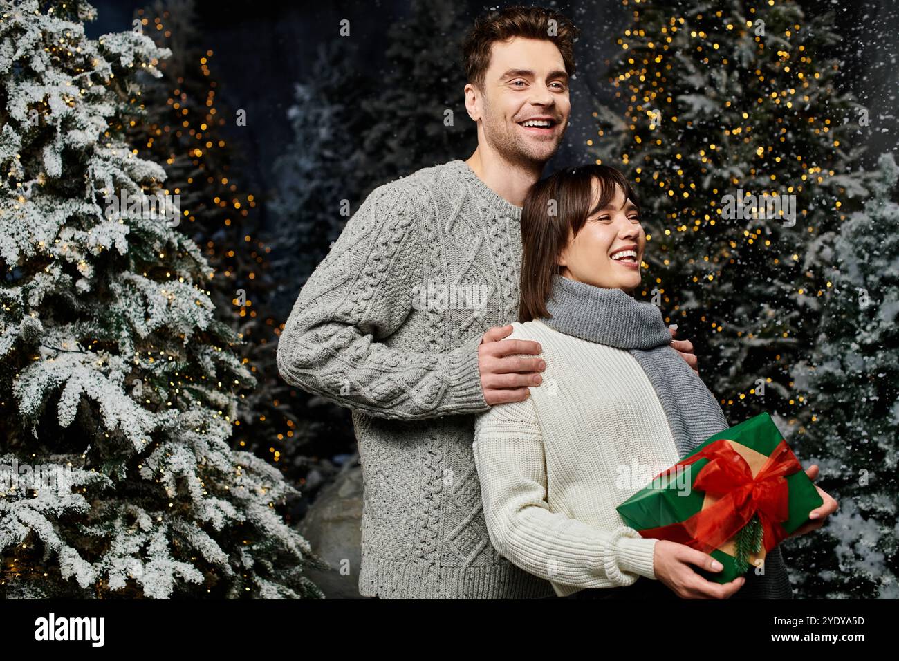 Questa deliziosa coppia abbraccia lo spirito natalizio, circondato da abeti innevati e decorazioni festose. Foto Stock