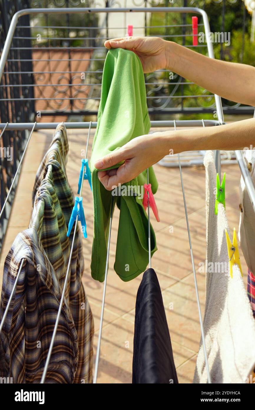 Le mani femminili tolgono abiti puliti asciugati al sole. Svolgere le normali faccende domestiche: Donna che tiene in mano una maglietta verde tolta dalla griglia di asciugatura Foto Stock