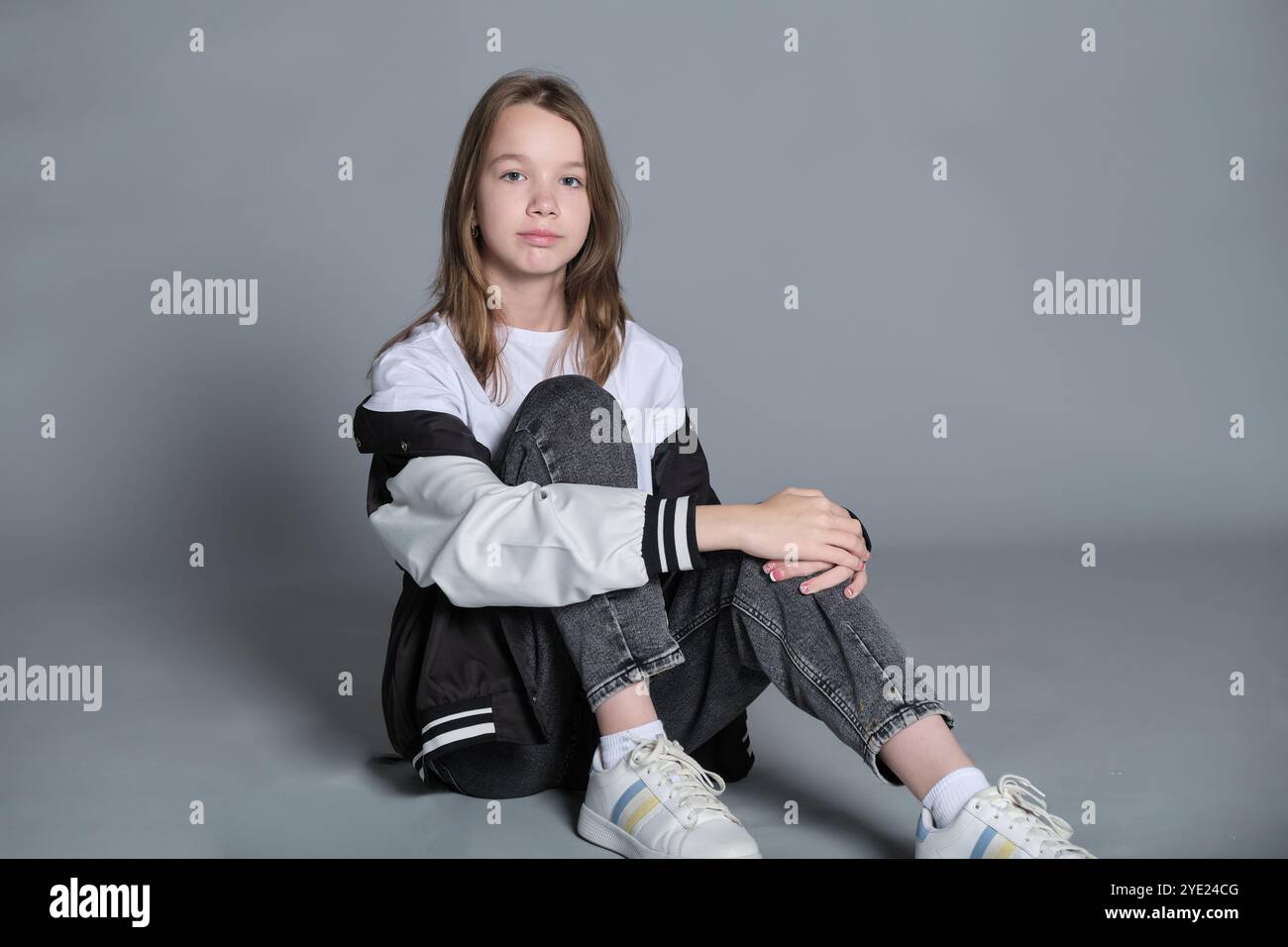 Una giovane ragazza siede a gambe incrociate sul pavimento, guardando attentamente di lato. Questa immagine cattura un momento sereno, riflettendo la natura introspettiva Foto Stock