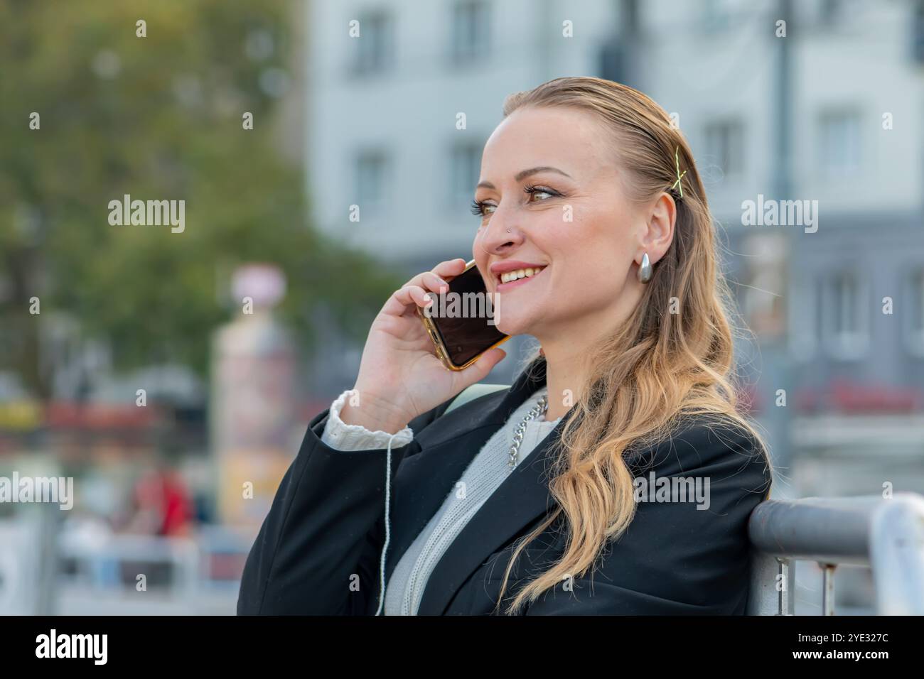 Una donna di mezza età di 40-45 anni che parla al telefono mentre si siede su una panchina in una città vivace, trascorrendo una piacevole giornata con un sorriso sul viso. Foto Stock