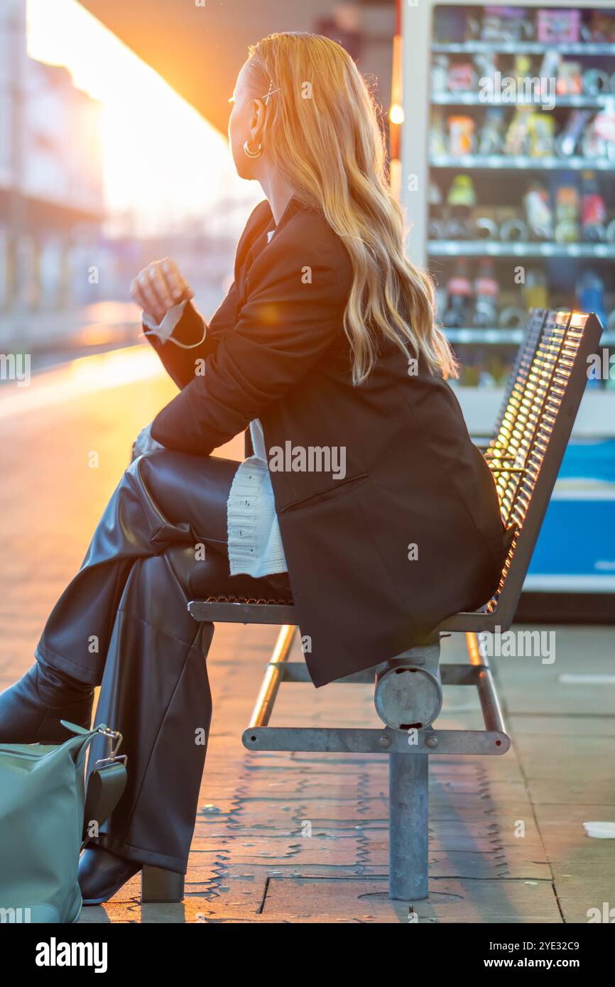 Una giovane donna aspetta in una stazione ferroviaria mentre il sole tramonta, emettendo una luce calda e creando un'atmosfera tranquilla durante gli spostamenti serali Foto Stock