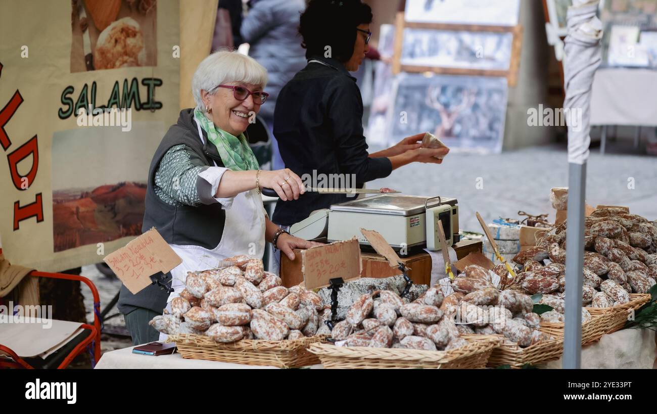 Un allegro venditore presenta una serie di salumi fatti in casa in un vivace mercato di Alba, Italia, coinvolgendo i clienti sotto la luce del sole Foto Stock