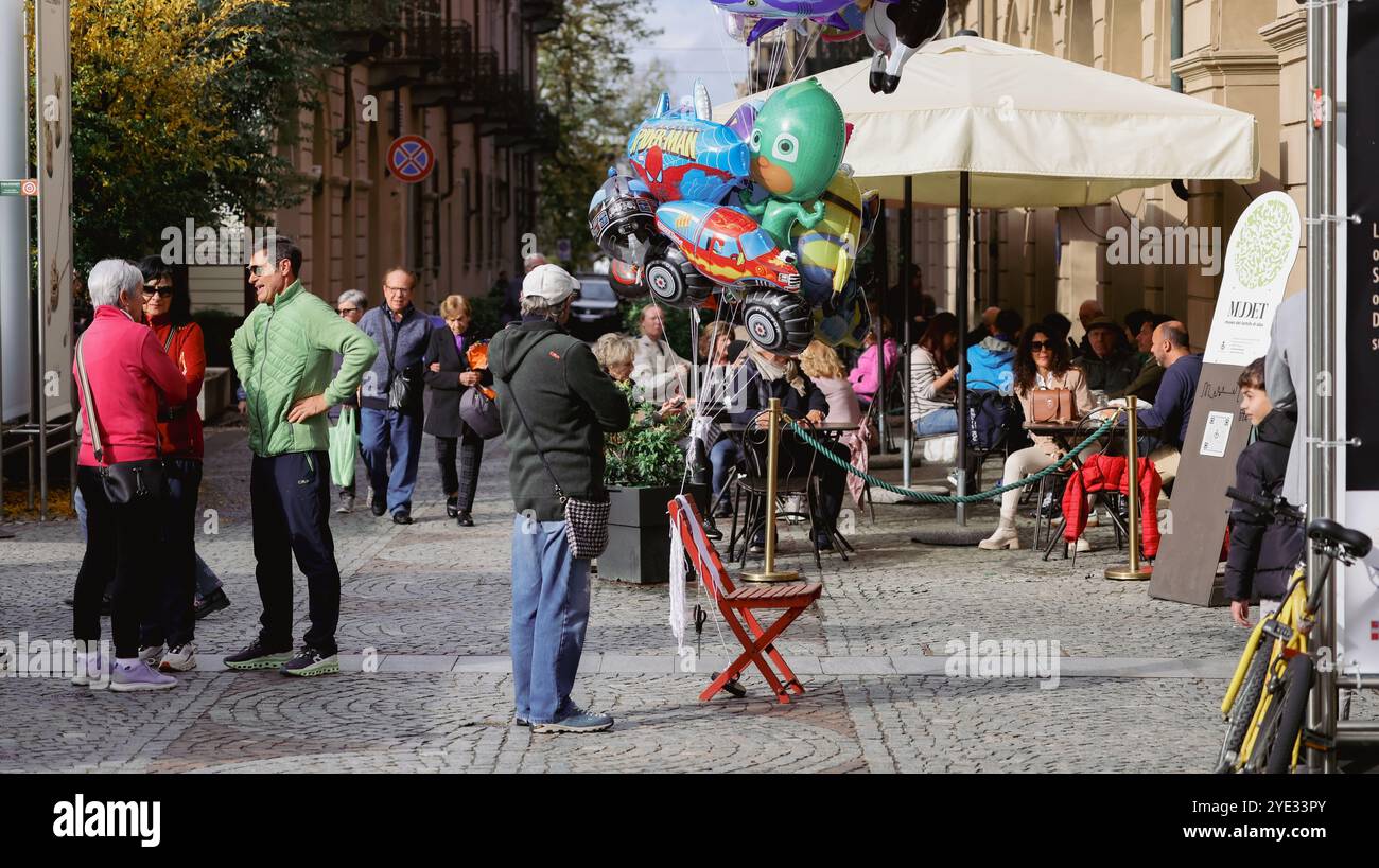 La gente si riunisce in una vivace strada di Alba, in Italia, godendosi una piacevole giornata. Palloncini colorati aggiungono allegria, mentre un po' di relax ai tavoli all'aperto Foto Stock
