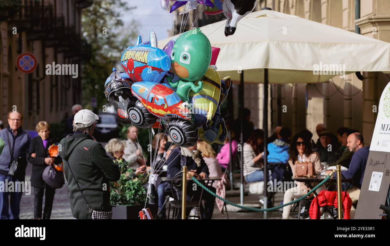 I palloncini dai colori vivaci fluttuano sopra una vivace strada di Alba, in Italia, dove le persone si godono l'atmosfera e socializzano in una giornata di sole Foto Stock