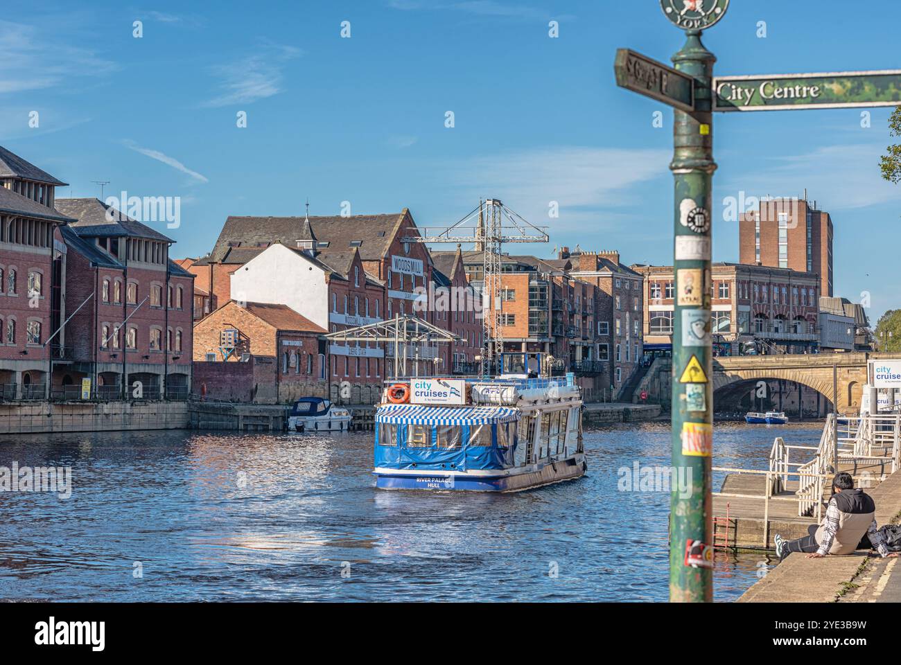 Un tour in barca scivola lungo un fiume con magazzini convertiti sulla riva lontana. Un ponte è in lontananza e un cartello è in primo piano. Foto Stock