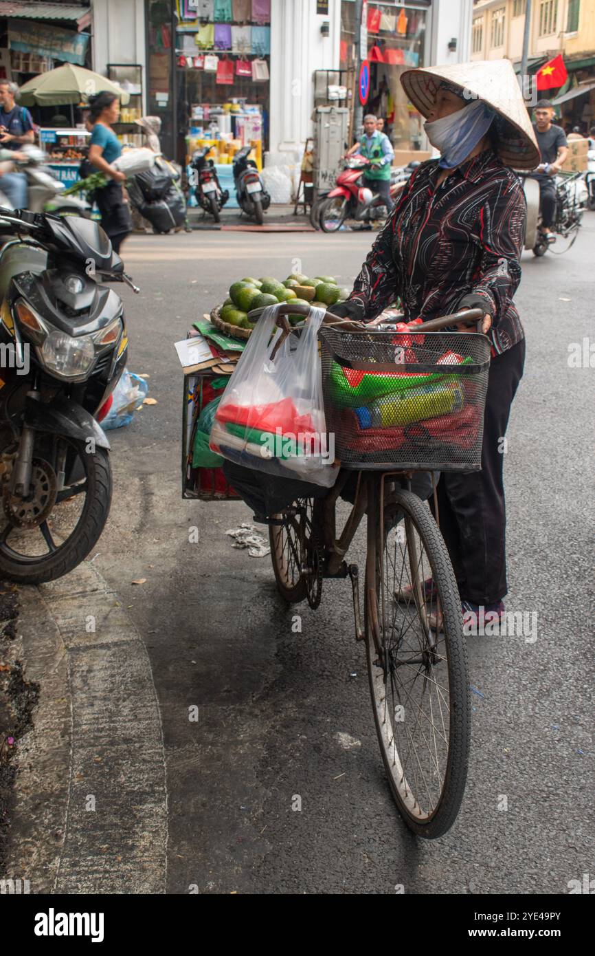 Hanoi, Vietnam: Una donna vietnamita che indossa il tradizionale cappello conico non la e che vende frutta porta la merce sulla sua bicicletta nel traffico cittadino Foto Stock