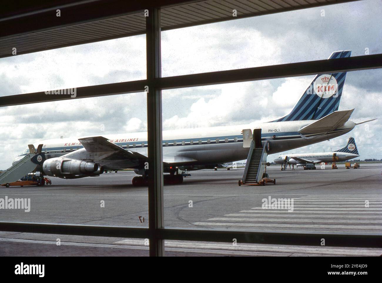 Amsterdam, Paesi Bassi. 1961 agosto – Una vista di un aereo di linea Douglas DC-8-53 della KLM (Royal Dutch Airlines) parcheggiato sul piazzale dell'aeroporto di Schiphol, Amsterdam. La fotografia, scattata attraverso una vedova, dall'interno di un edificio del terminal, mostra l'aeromobile in fase di preparazione per il volo. Questo aereo è stato consegnato alla KLM nel maggio 1961, solo tre mesi prima di questa fotografia, ed è mostrato nella sua livrea originale. Rimase in servizio con la compagnia fino al 1976. Visibile in lontananza è un aereo di linea KLM Convair CV-340. Foto Stock