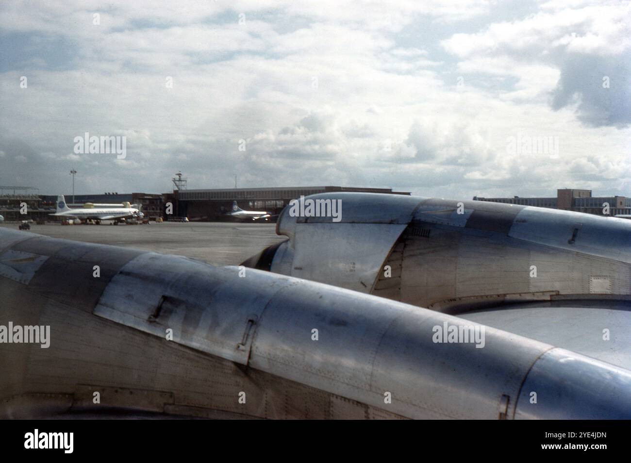 Amsterdam, Paesi Bassi. 1961 agosto – Una vista sull'ala di un aereo di linea Lockheed L-188 Electra, con i suoi motori in funzione. L'aereo si trova sul piazzale dell'aeroporto Schiphol di Amsterdam. Gli hangar e gli edifici terminal sono visibili in lontananza, insieme agli aerei di linea KLM Convair CV-340 e Pan American World Airways Douglas DC-7. Foto Stock
