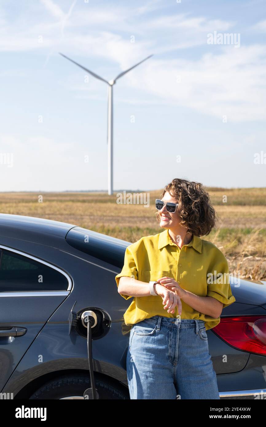 Donna con capelli curvilinei che indossa camicia gialla e occhiali da sole si trova accanto alla ricarica di un'auto elettrica. Turbine eoliche sullo sfondo. Foto Stock