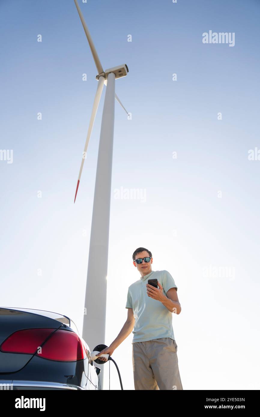 Uomo con auto elettrica in carica. Turbina eolica sullo sfondo. Foto Stock