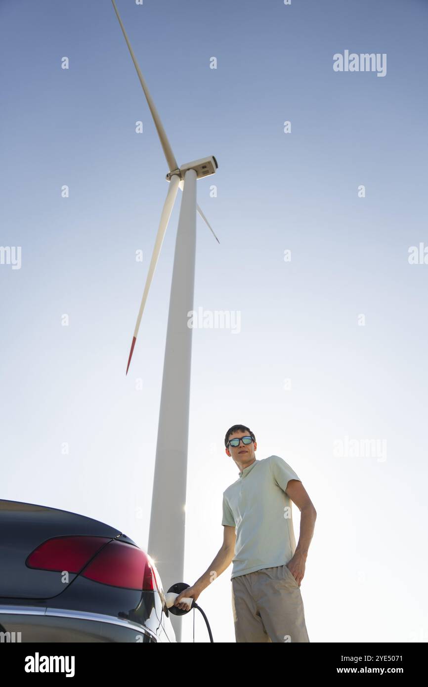 Uomo con auto elettrica in carica. Turbina eolica sullo sfondo. Foto Stock