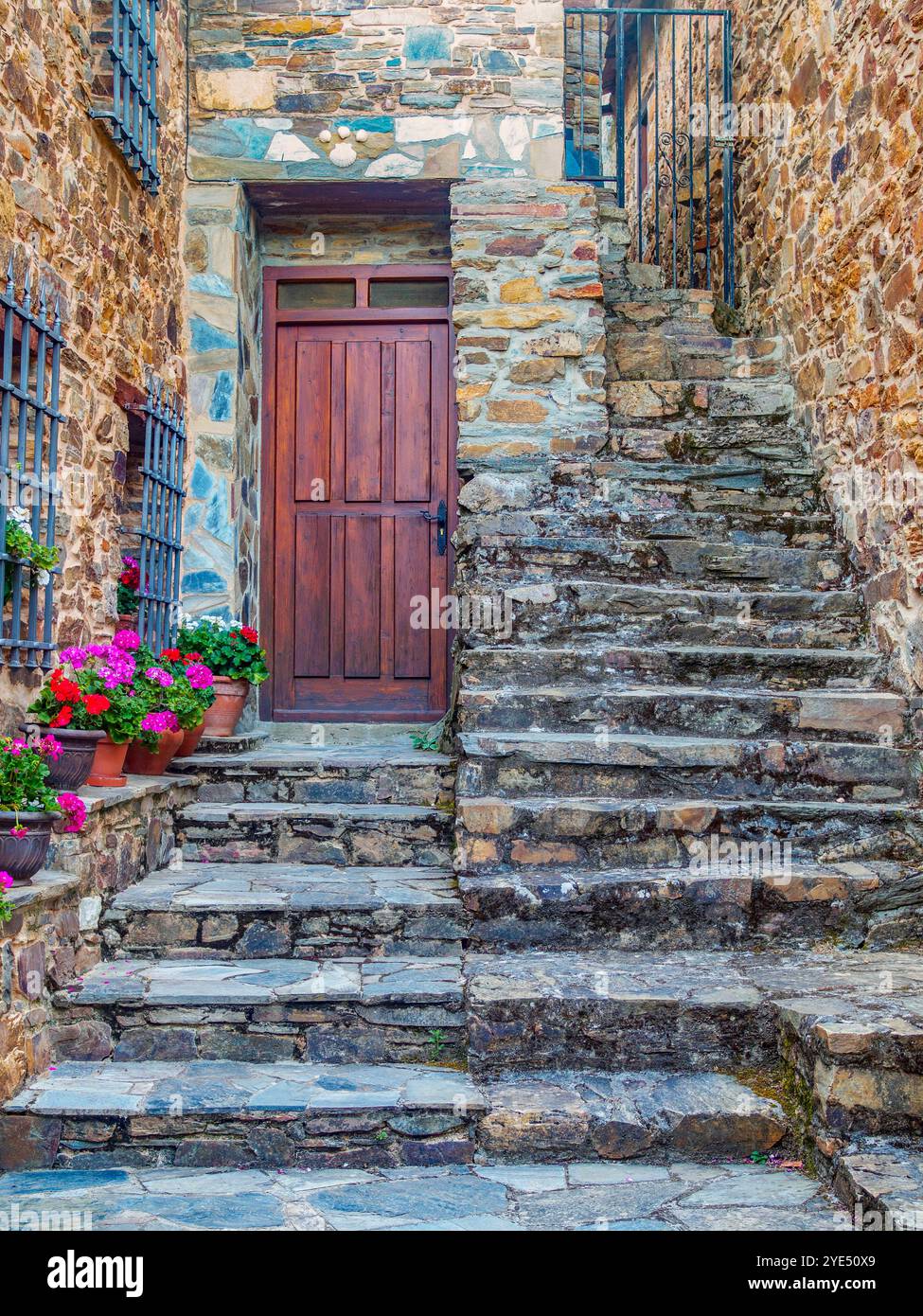 Affascinante porta in legno adagiata su una facciata in pietra intemprata, fiancheggiata da colorati fiori in vaso. Antichi gradini in pietra conducono all'ingresso. Foto Stock