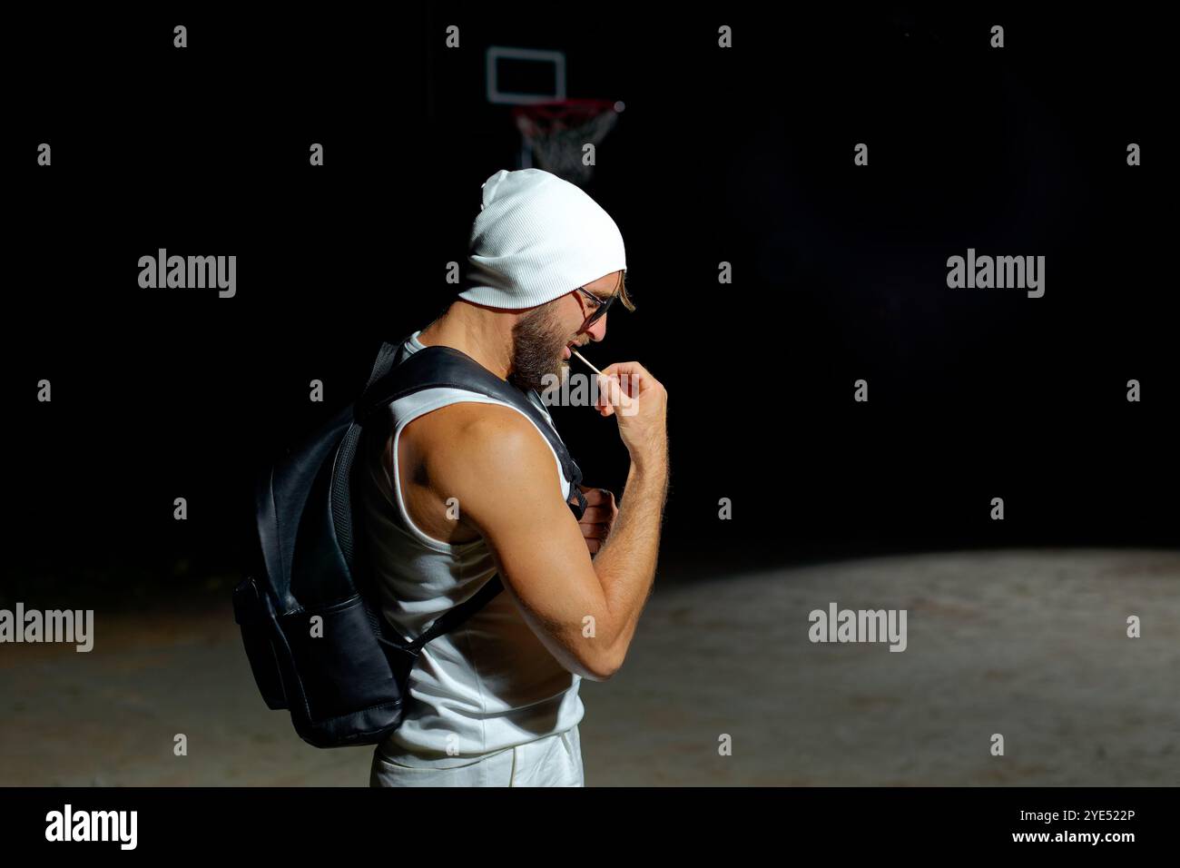 Un individuo muscoloso si posa in un'illuminazione soffusa, indossando occhiali da sole e un berretto mentre tiene in mano lo zaino. La sua posizione sicura riflette una potenza e uno stile Foto Stock