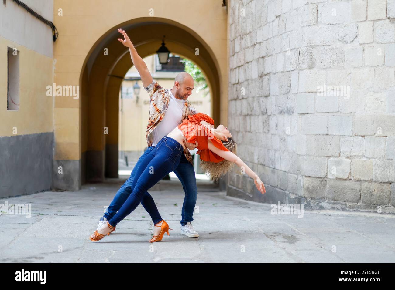 Una coppia esegue una energica routine di danza in un vicolo stretto, con l'uomo che sostiene la donna in un tuffo drammatico, evidenziando la loro passione e sk Foto Stock Una coppia esegue una energica routine di danza in un vicolo stretto, con l'uomo che sostiene la donna in un tuffo drammatico, evidenziando la loro passione e sk Foto Stock