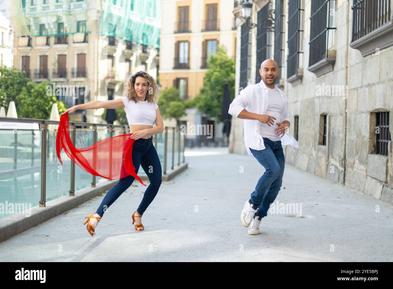 Una coppia energica si esibisce in danza latina su una strada soleggiata della città. La donna gira una sciarpa rossa mentre entrambe mostrano espressioni gioiose, abbracciando il vib Foto Stock