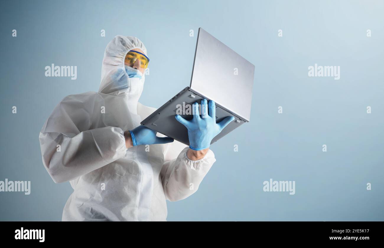 Lavoratore di laboratorio, un medico professionista in una tuta protettiva, guanti e occhiali di protezione, utilizza un computer portatile su uno sfondo blu Foto Stock