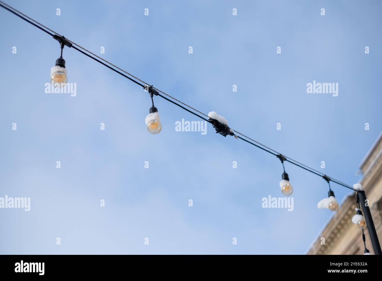 La neve poggia su luci decorative a corda appese sopra uno spazio all'aperto, adagiate su un cielo blu brillante, creando un'atmosfera invernale accogliente. Foto Stock
