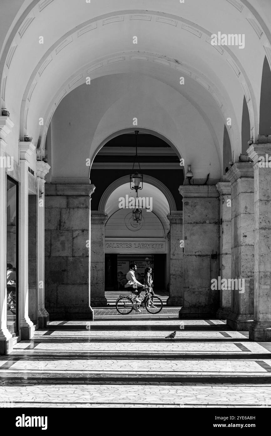 Immagine in bianco e nero di un ciclista che cavalca gli archi in Place Massena, una piazza storica di Nizza, Francia Foto Stock