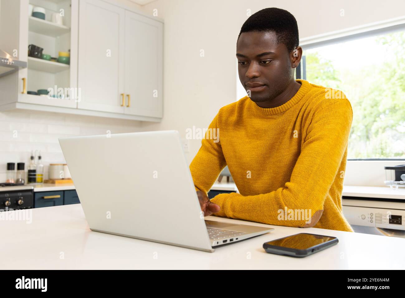 Uomo afroamericano in cucina che usa un computer portatile, indossa un maglione giallo, uno smartphone sul bancone, a casa Foto Stock
