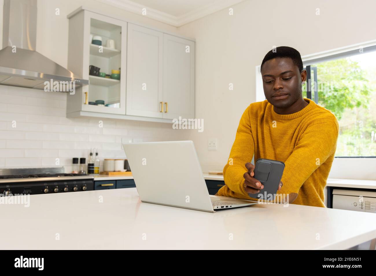A casa, un giovane afroamericano che usa smartphone e computer portatile in una cucina moderna Foto Stock