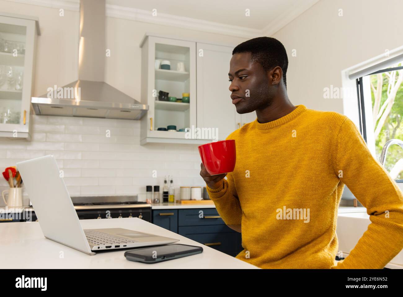 Uomo afroamericano in cucina che beve caffè mentre usa il computer portatile, concentrandosi sullo schermo, a casa Foto Stock
