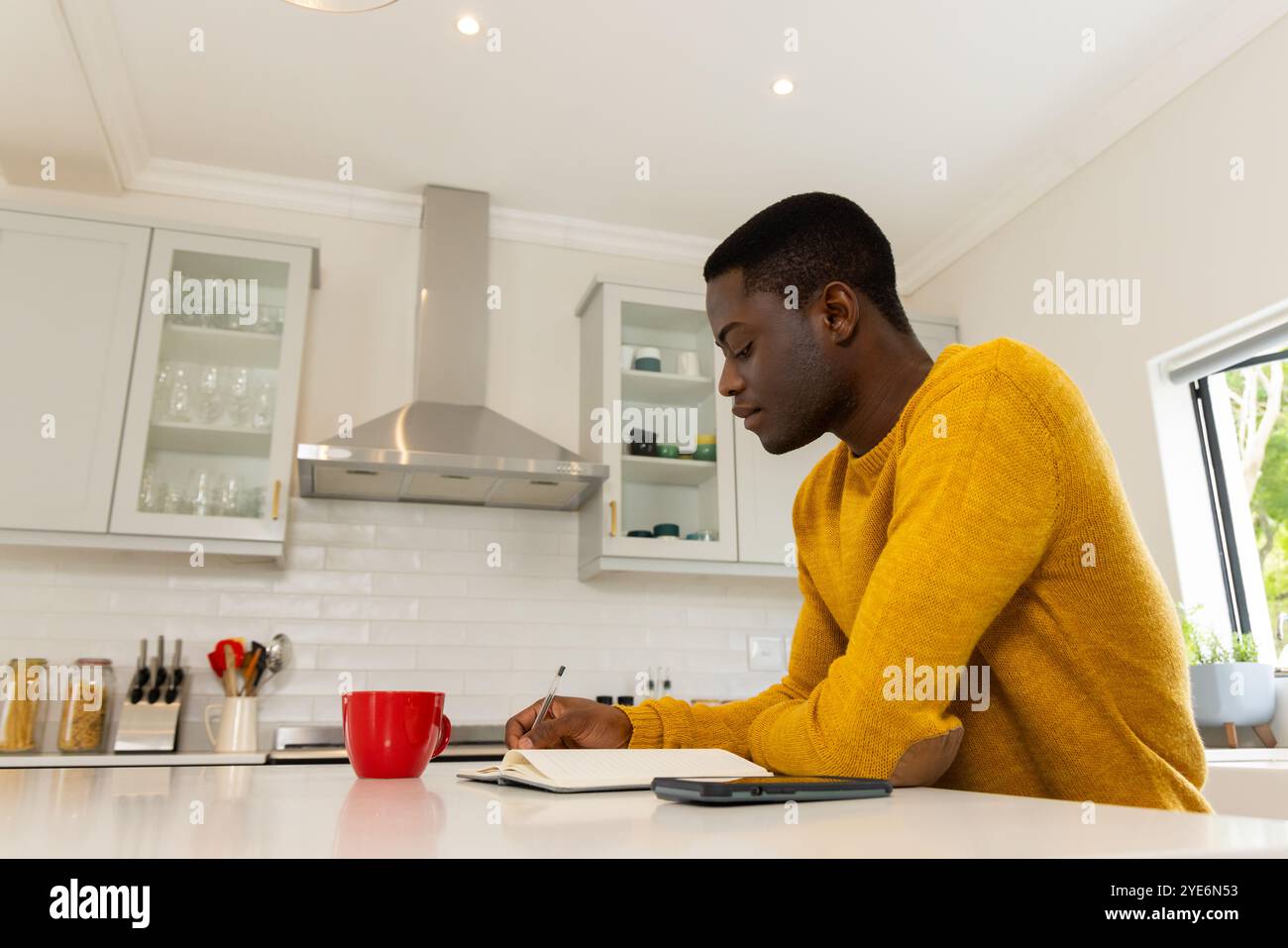 Uomo afroamericano in cucina che scrive su un notebook con smartphone e tazza rossa nelle vicinanze, a casa Foto Stock