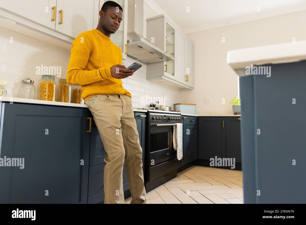 Uomo afroamericano in cucina che usa lo smartphone, indossa un maglione giallo, a casa Foto Stock