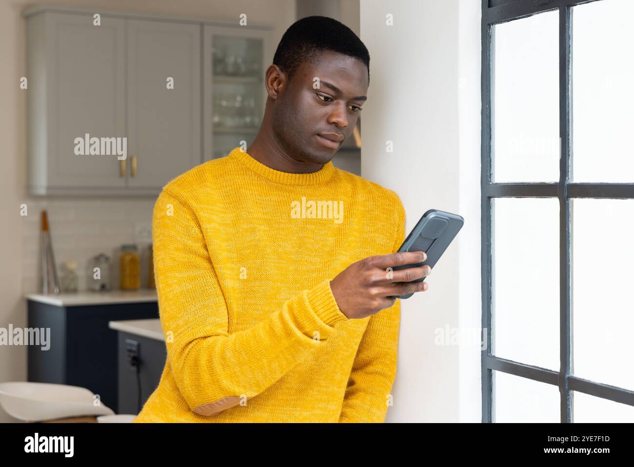 Uomo afroamericano in maglione giallo che usa lo smartphone vicino alla finestra a casa, che sembra concentrato Foto Stock