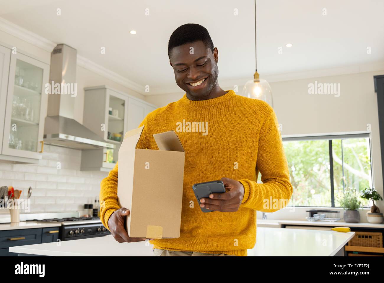 Sorridente afroamericano in cucina che controlla lo smartphone mentre apri la cassetta di consegna, a casa Foto Stock