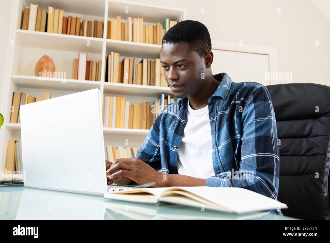 Uomo afroamericano concentrato che usa un computer portatile a casa con un libro aperto nelle vicinanze Foto Stock