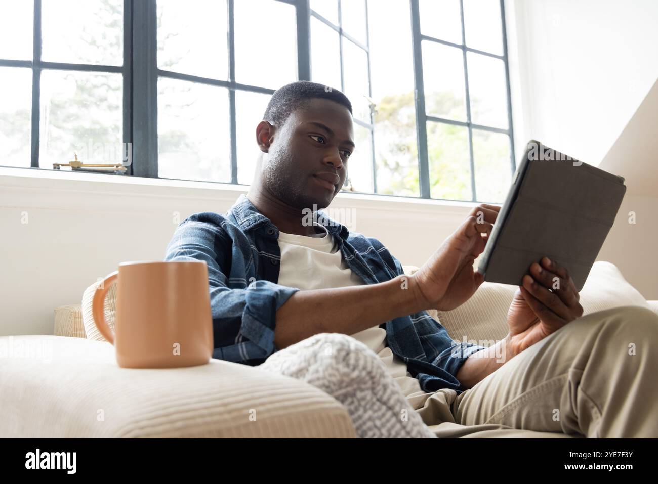 Rilassatevi a casa, un uomo afroamericano che usa un tablet con una tazza di caffè sul comodo divano Foto Stock