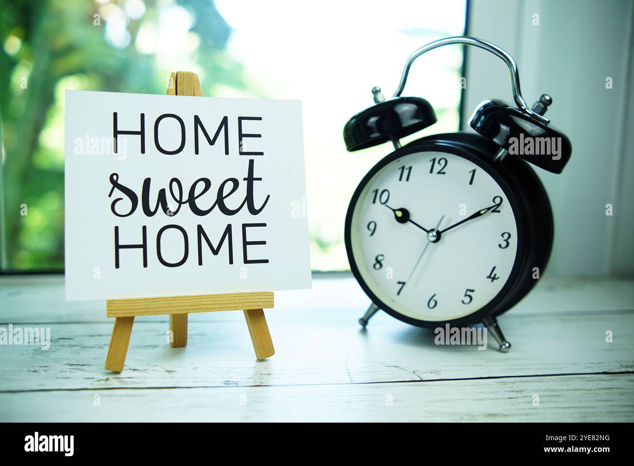 Home Sweet Home cita il testo su carta con cavalletto e sveglia sullo sfondo della finestra Foto Stock