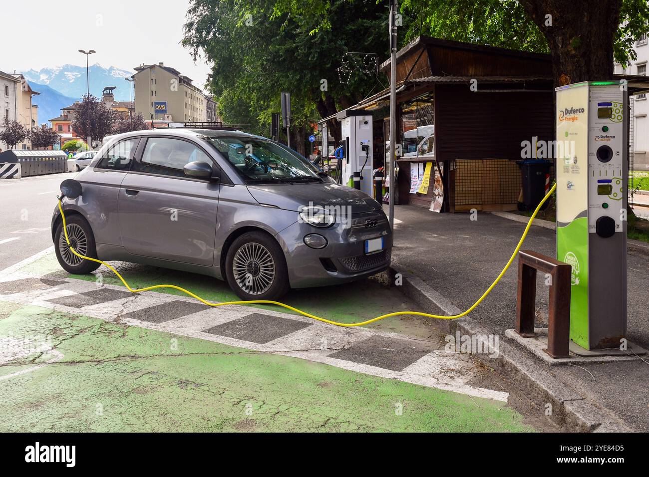 Una Fiat 500 elettrica in una stazione di ricarica nella centralissima Piazza della Repubblica, con il Monte bianco sullo sfondo, Aosta, Valle d'Aosta, IT Foto Stock
