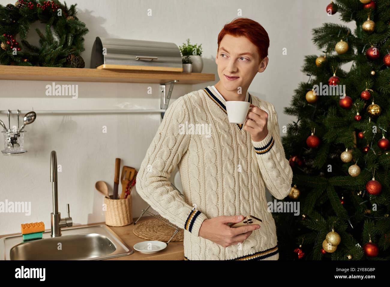 Un giovane tiene una tazza, assaporando un momento di gioia accanto a un albero di Natale nella sua cucina. Foto Stock