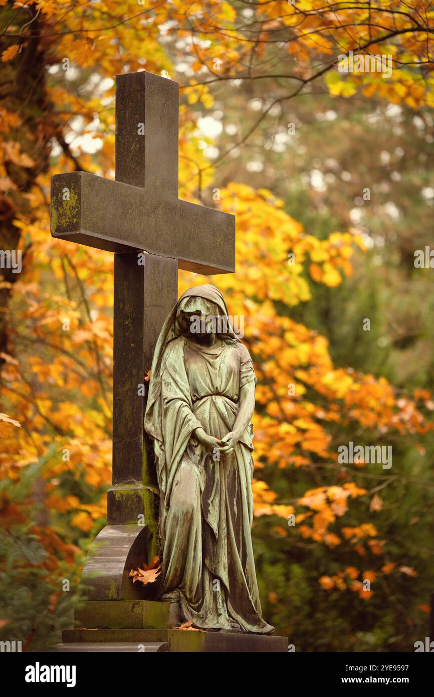 una statua femminile del cimitero con il viso nero proveniente dagli agenti atmosferici si appoggia con le mani piegate contro una croce di fronte alle scintillanti foglie autunnali nel backgrou Foto Stock